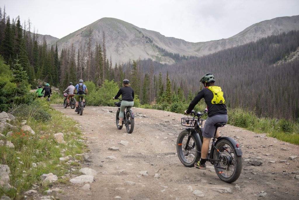 Six e-bike riders on a dirt road with mountain peaks in the background