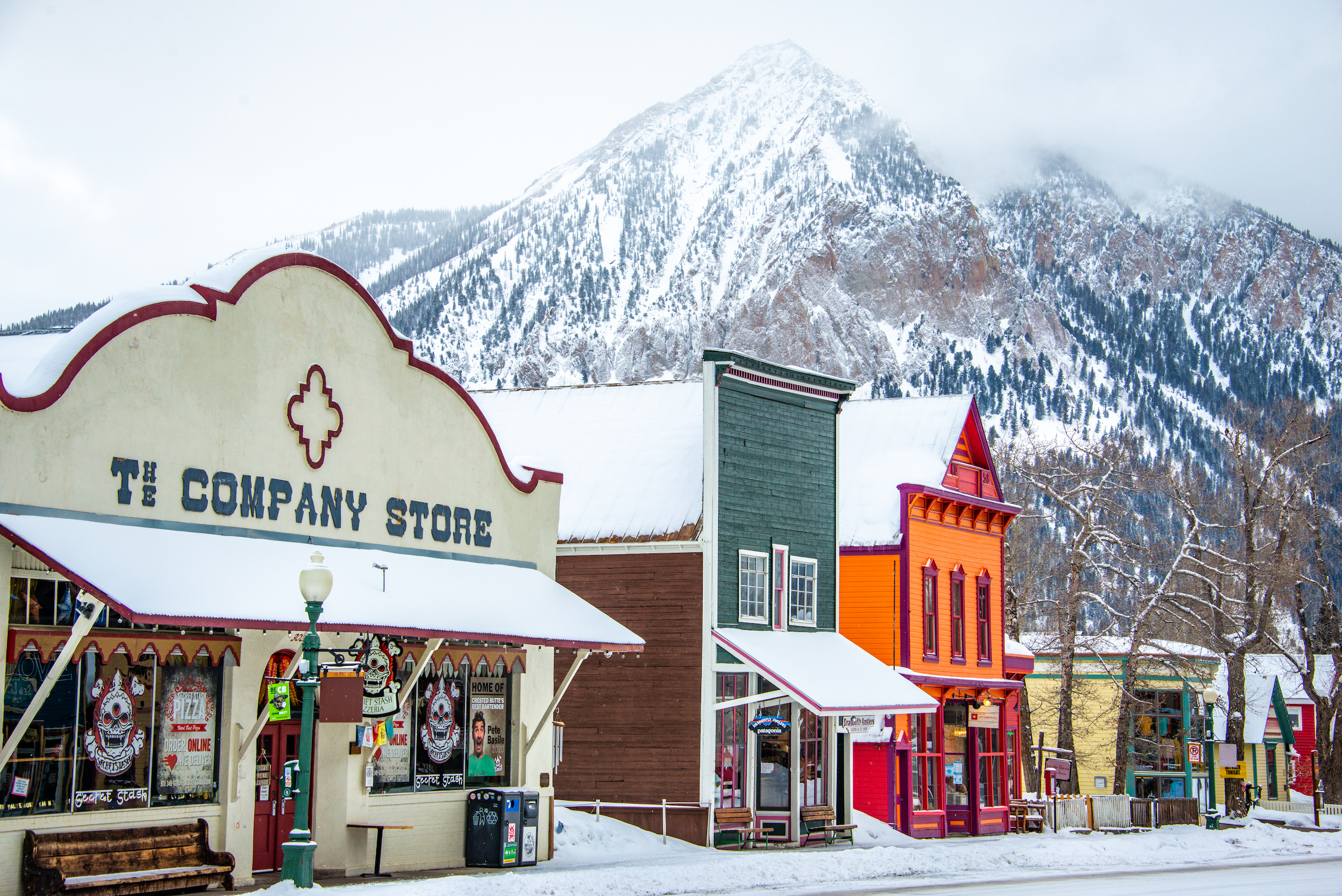 A downtown street covered in snow with a snowcapped mountain peak in the background. This is Elk Avenue in winter in Crested Butte
