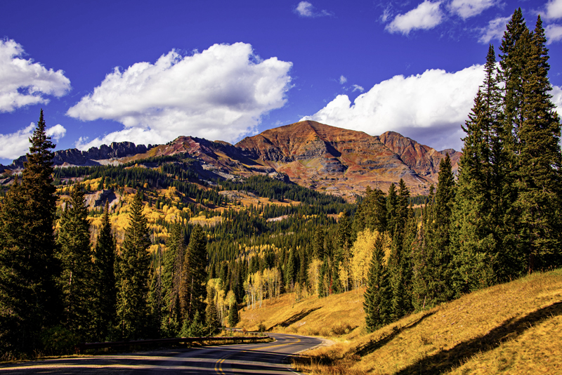 Crested Butte fall hiking photo of a huge red mountain with green spruce trees and yellow aspens in the foreground. A paved road winds through the trees.