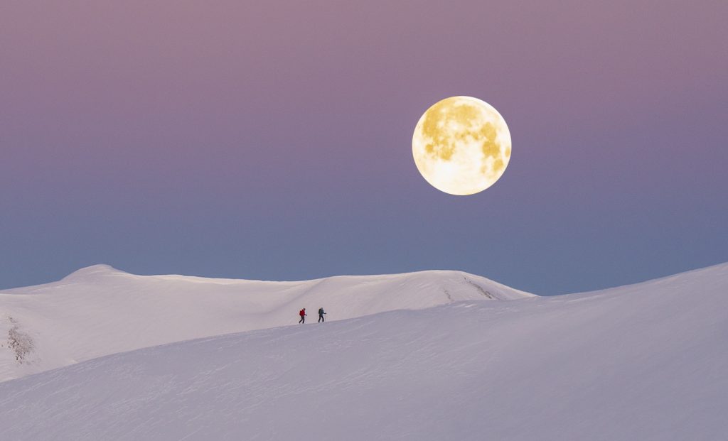 Two skiers cross a ridge covered in snow with a full moon rising against a purple sky
