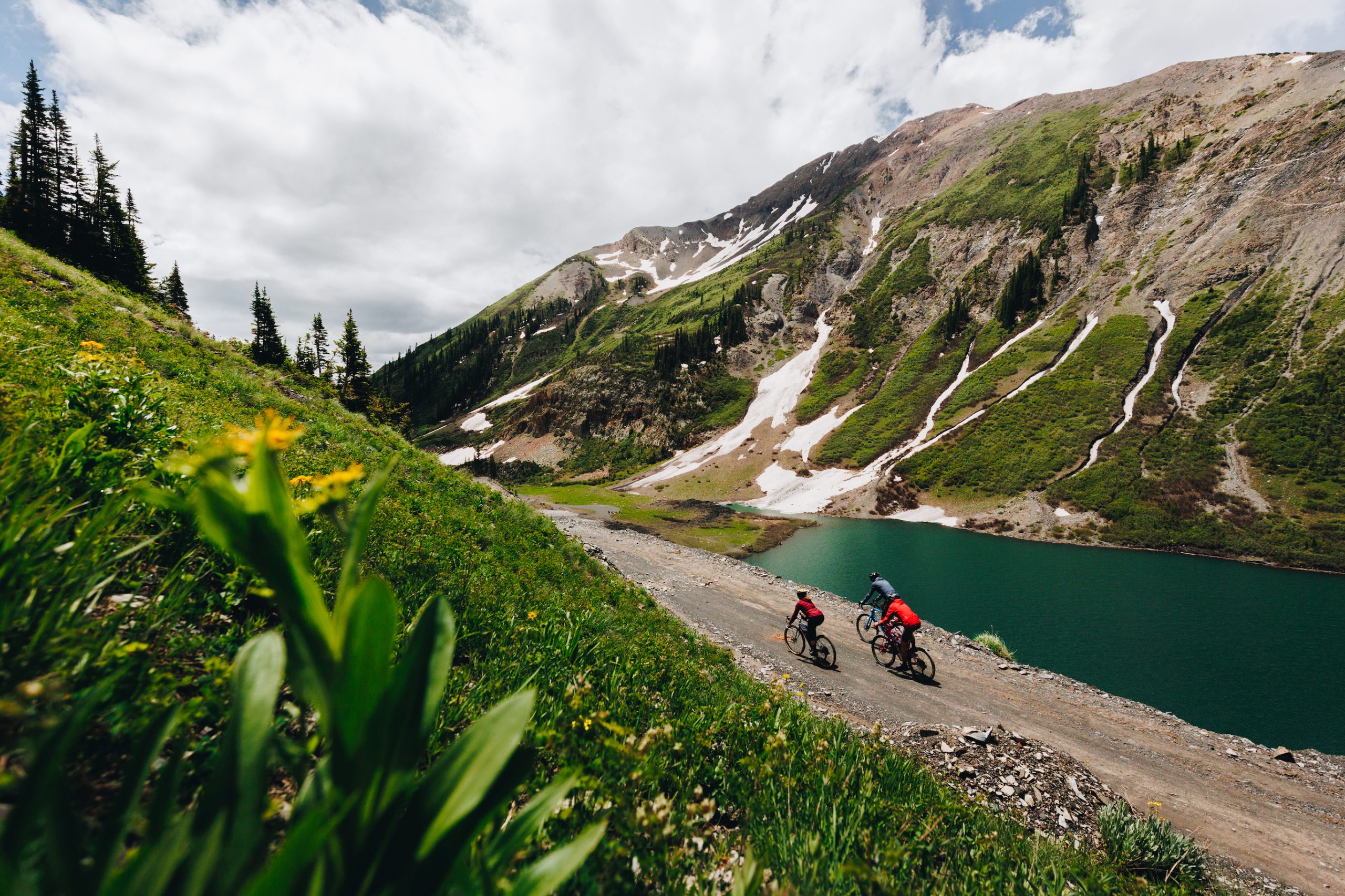 Three people ride bikes on a dirt road next to a lake