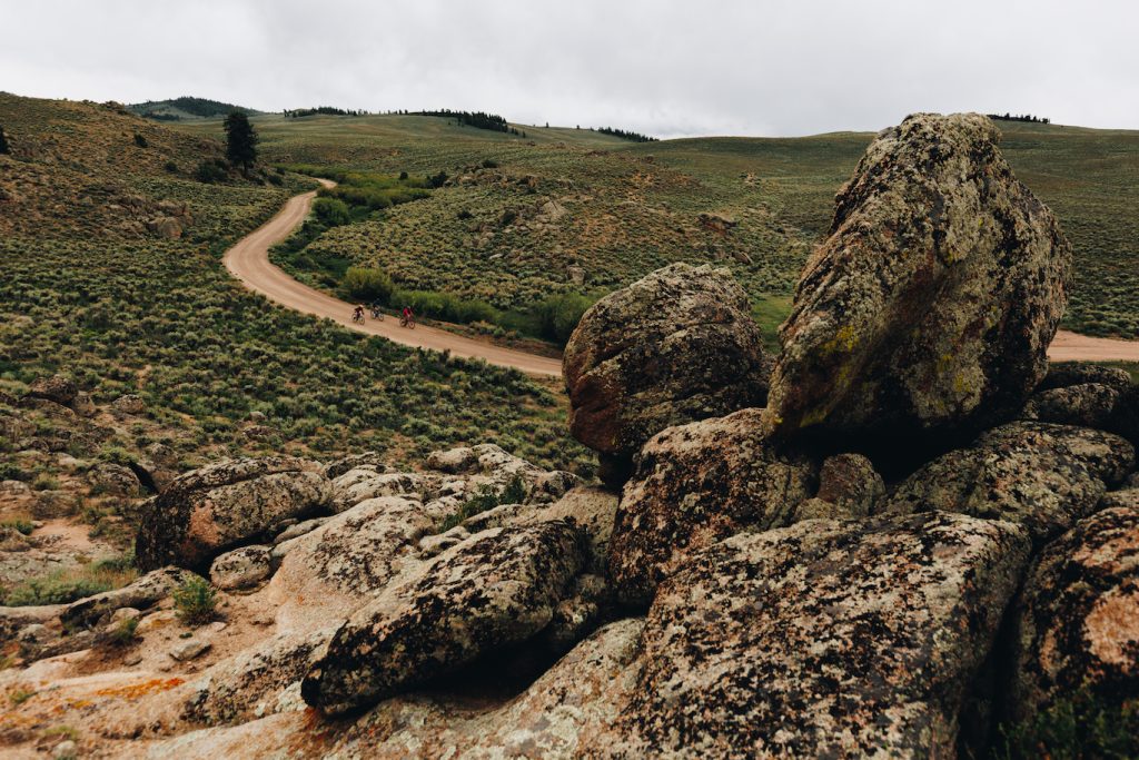 Three cyclists ride a curving gravel road through a field of green sage brush around a large rock formation 