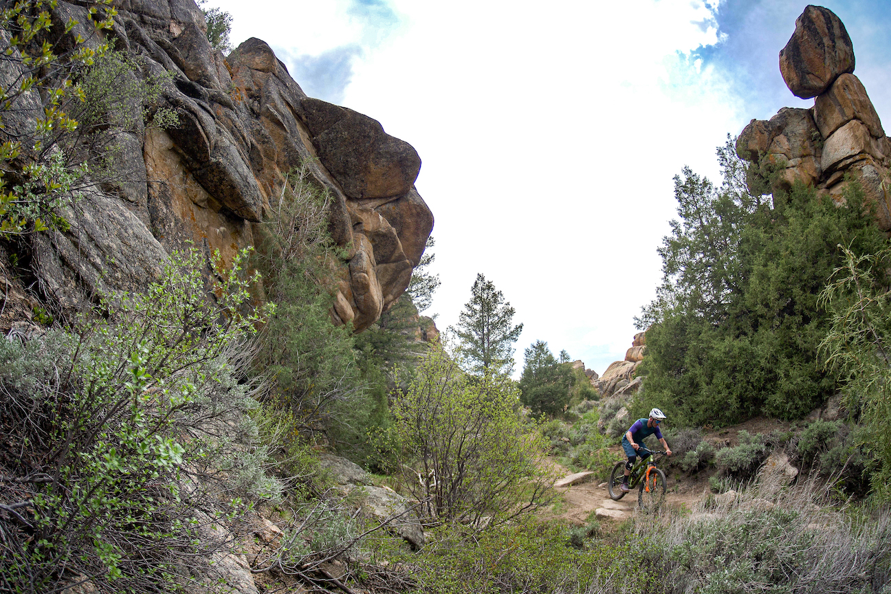 A mountain biker rides on a trail nestled between rock faces and trees and bushes.