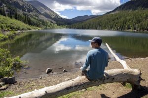 A man sits on a log on the shore of Mirror Lake in Tincup, Colorado on a summer day.