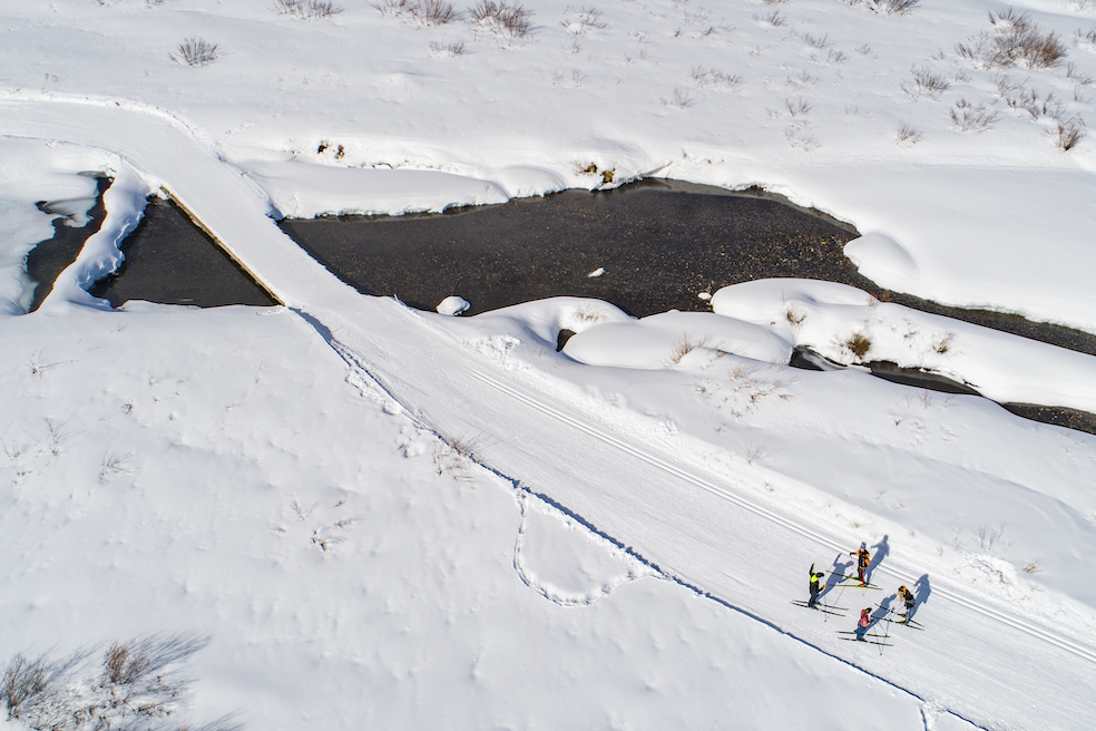 An aerial view of a Nordic ski track, which is corduroy like snow with a parallel track. The track goes over a stream and there are four skiers in the bottom right corner 
