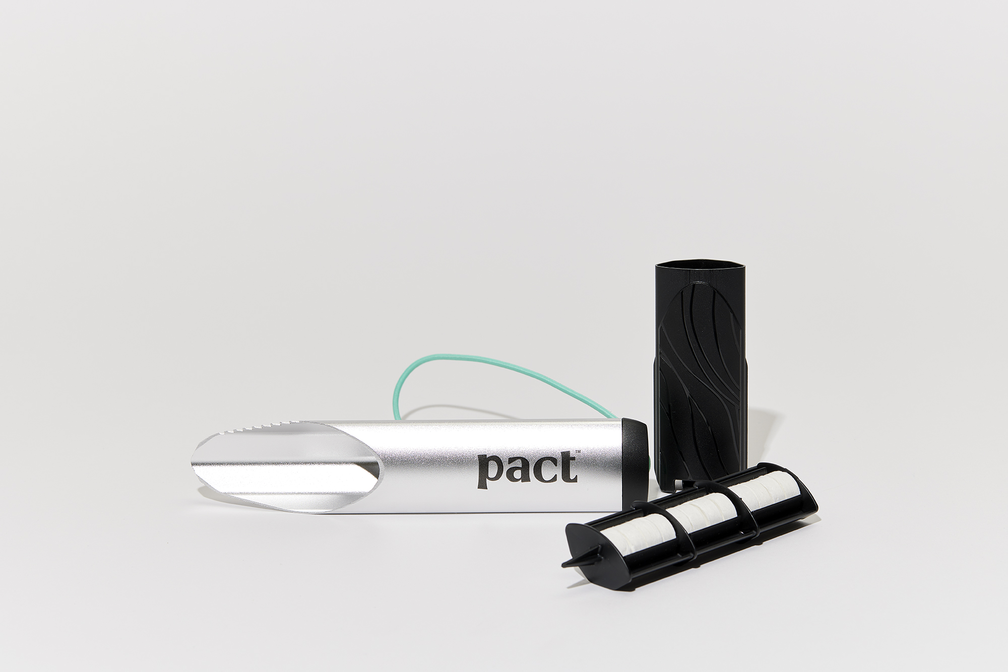 A small silver shovel that says "pact." It is serrated on one side. There is also a black container that holds white round compressed wipes and a black rectangular pouch.