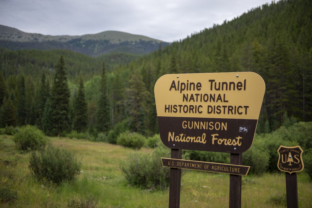 A yellow and brown sign with the words "Alpine Tunnel National Historic District. Gunnison National Forest" in front of a forest and mountain view.