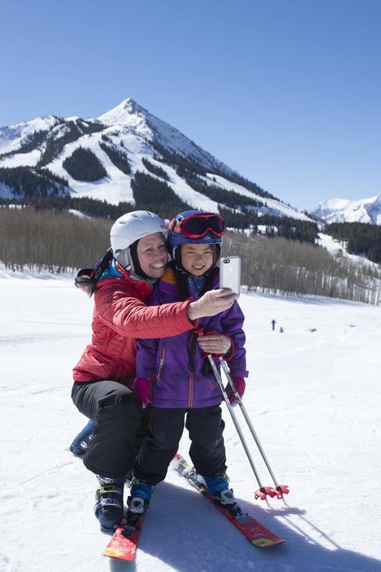 Ski with kids at Crested Butte. Kids can learn how to ski on the Peachtree Lift, take a lesson or shred with the adults. A woman on skis takes a selfie with a kid on skis in front of a pointy mountain peak. 