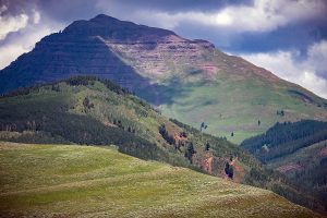 A massive, rocky peak, Teocalli Mountain, looms over a green alpine landscape in Colorado.