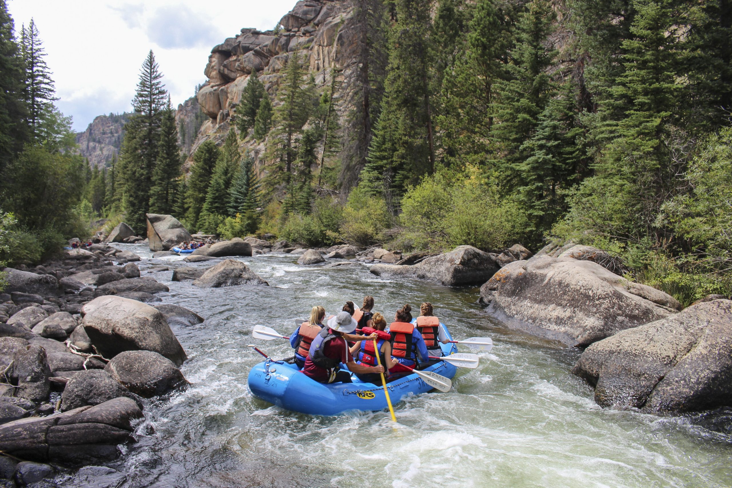 A group of seven people sit in an inflatable raft on a whitewater river with rocks